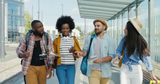 Multietniska män och kvinnor turister går tillsammans vid busshållplatsen och talar glatt på sommardagen. Grupp av blandade raser snygga glada män och kvinnor resenärer promenader och ha kul. — Stockvideo