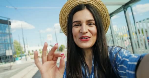 Gros plan de jeune touriste heureuse caucasienne en chapeau parlant, agitant la main et souriant à la caméra en plein air en été. POV de jolie voyageuse ayant vidéochat. Concept de vidéochat. — Video