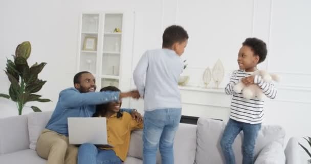 Cute small African American kids, boy and girl playing and jumping on couch in living room. Parents smiling and sitting on sofa with laptop computer. Children having fun at home. Mother and father. — Stock Video