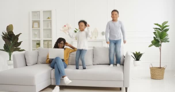 Lindos niños afroamericanos pequeños, niños y niñas jugando y saltando en el sofá en la sala de estar. Hermosa madre trabajando en el ordenador portátil y sentado en el sofá al lado. Feliz mamá y niños ruidosos. — Vídeos de Stock