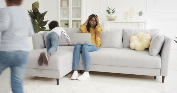 Müde verzweifelte afroamerikanische Mutter sitzt auf der Couch eines Hauses mit Laptop-Computer und hält ihren Kopf mit Händen. Laute, schlecht gelaunte Kinder, die herumrennen, schreien und auf Sofa springen. — Stockvideo