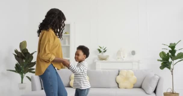 Feliz alegre mãe afro-americana dançando e se divertindo com a filha na sala de estar em casa durante o confinamento. Mamãe dança com pequeno garoto bonito e rindo. Pequena criança alegre curtindo música. — Vídeo de Stock