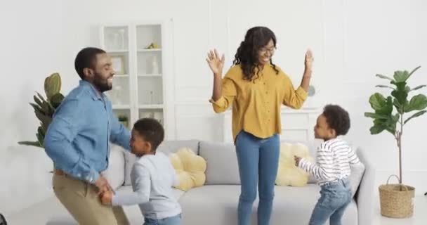 Feliz alegre familia afroamericana bailando y divirtiéndose en la sala de estar en casa durante el encierro. Madre y padre bailan con su pequeño y lindo hijo y su hija. Niños pequeños disfrutando de la música con sus padres. — Vídeos de Stock