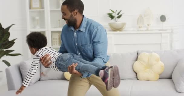 Fröhlich fröhlicher afroamerikanischer Vater, der mit seiner kleinen süßen Tochter im Wohnzimmer spielt. Papa hält ein kleines Kind und bewegt es wie ein Flugzeug, das zu Hause in der Luft fliegt. Eltern haben Spaß mit Kind. — Stockvideo