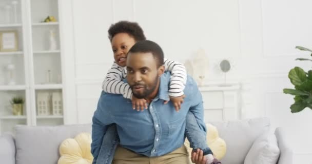 Vrolijke gelukkige jonge Afro-Amerikaanse vader houdt zijn schattige dochtertje achter de rug en wervelt in een gezellige woonkamer. Papa speelt thuis met een kind. Mooi klein meisje lachen en plezier hebben. — Stockvideo