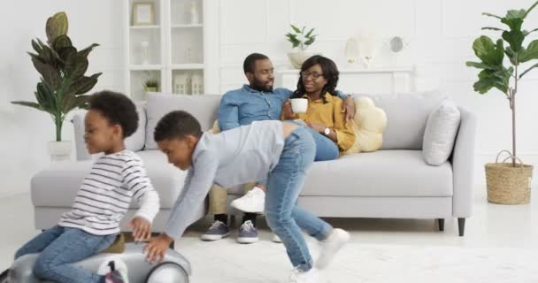 Lindos niños afroamericanos pequeños jugando en la sala de estar. Madre y padre sentados en abrazos con café en el sofá en el fondo. Pequeño niño montando hermana en coche de juguete. En casa. Familia juntos. — Vídeos de Stock