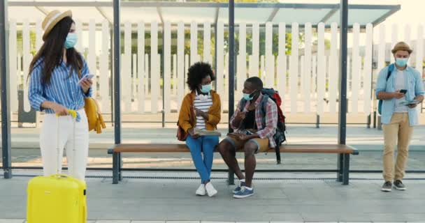 Jeunes hommes et femmes de races mixtes portant des masques médicaux debout à l'arrêt de bus et attendant le transport. Touristes multiethniques hommes et femmes à la gare. Concept de voyage. — Video