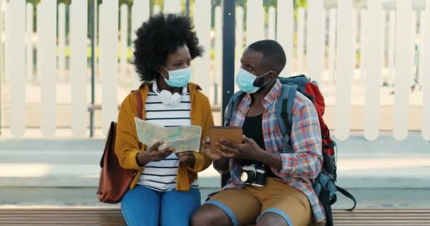 Afro-Amerikaanse jonge paar toeristen in medische maskers zitten op de bank bij de bushalte en wachten op vervoer. Mooie vrouw met stadsplattegrond en knappe man met tablet op zoek naar route. — Stockvideo