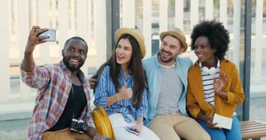 Karışık ırklar genç turistler, şık erkek ve kadınlar otobüs durağında bankta oturuyor ve cep telefonu kamerasıyla selfie çekiyorlar. Güzel dişiler ve yakışıklı erkekler nakil için bekliyor ve fotoğraf çekiyorlar.