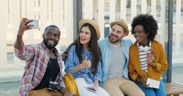 Giovani turisti di razze miste, uomini e donne alla moda seduti sulla panchina alla fermata dell'autobus e scattare foto di selfie sulla fotocamera del telefono cellulare. Belle femmine e bei maschi in attesa di trasporto e fare foto — Video Stock