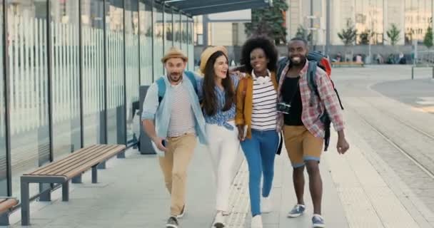 Unga glada kaukasiska och afroamerikanska turister har roligt utomhus, springa och hoppa. Glada glada blandraser män och kvinnor reser tillsammans. I staden vid busshållplatsen. Multietniskt. — Stockvideo