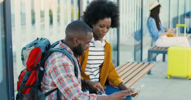 アフリカ系アメリカ人の若いカップルの観光客のバス停留所でベンチに座って、話をし、輸送を待っている。街の地図とハンサムな男とかなりの女性の方法と計画ルートを探しています。旅行者. — ストック動画