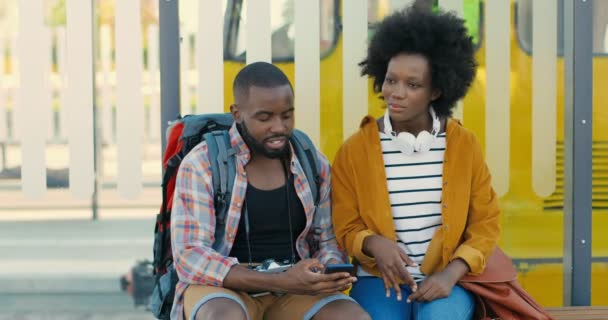 Afro-americanos jovem casal de turistas sentados no banco na parada de ônibus, conversando e esperando por transporte. Mulher bonita e homem bonito à procura de maneira no smartphone e rota de planejamento. Viajantes. — Vídeo de Stock