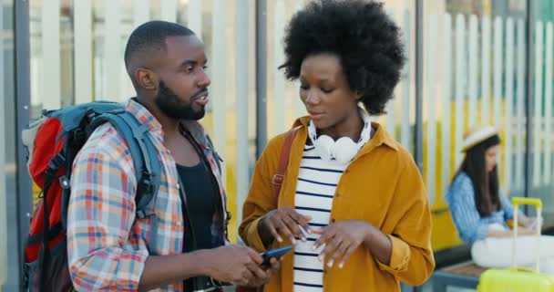 Mladí dobře vypadající Afroameričané stojí na autobusové zastávce a povídají si. Pohledný muž s mobilním telefonem a hezkou ženou mluví a čekají na transport. Cestující turisté. — Stock video