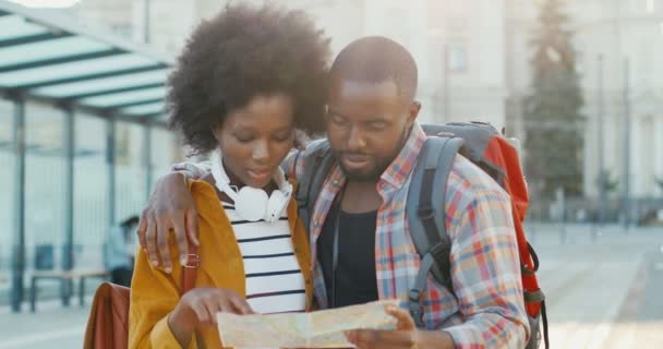 Portrét šťastného mladého afroamerického páru turistů stojících na ulici na autobusové zastávce a dívajících se na mapu. Krásná žena a pohledný muž plánuje trasu s plánem města. Koncept cestování. — Stock video