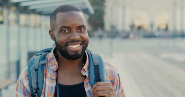 Portret van een knappe jonge, vrolijke Afro-Amerikaanse man met een rugzak die vrolijk naar de camera lacht. Close-up van mannelijke toerist glimlachte gezicht. Bij de bushalte. Vrolijke stijlvolle man reiziger op straat. — Stockvideo