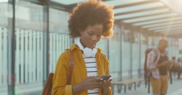 Joven hermosa mujer afroamericana mensaje de texto en el teléfono inteligente y sonriendo alegremente en la estación de tren. Atractivo viajero femenino tocando y desplazándose en el teléfono móvil en la parada de autobús. — Vídeo de stock