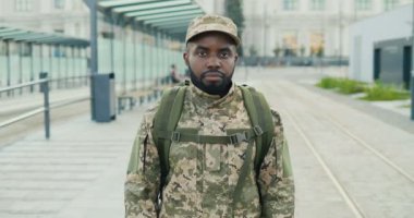 Ciddi bir Afro-Amerikan yakışıklı, şapkalı, sırt çantalı ve sokakta duran genç bir erkek askerin portresi. Asker bir adam tren istasyonunda kameraya bakıyor. Otobüs durağı. Askeri üniforma. Yakınlaştırılıyor.