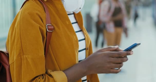 Primer plano del dispositivo de mano femenina afroamericana sosteniendo el teléfono inteligente y tocando en la pantalla. Al aire libre. Turista mujer desplazándose y escribiendo en el teléfono móvil en la calle en la parada de autobús. — Vídeos de Stock