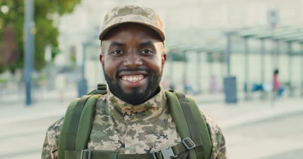 Ritratto di allegro afroamericano bel giovane soldato maschio in berretto con zaino sorridente per strada. Primo piano di militare maschile con sorriso felice alla stazione ferroviaria. Fermata dell'autobus. Uniforme militare. — Video Stock