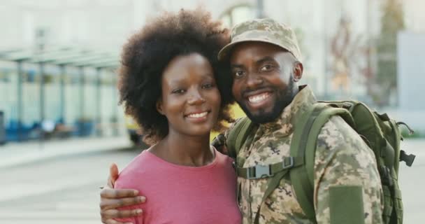Portrait of young cheerful African American couple hugging and smiling to camera outdoor in city. Handsome happy man soldier in military uniform hugs and embracing girlfriend. Meeting of guy and girl. — Stock Video