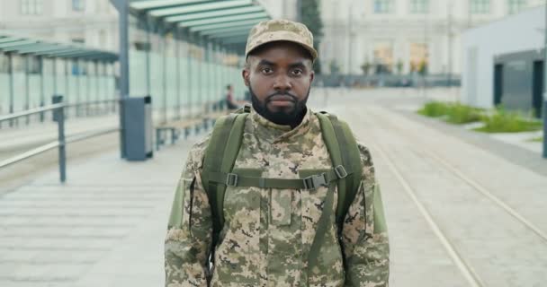 Portret van een Afro-Amerikaanse knappe jonge mannelijke soldaat in pet met rugzak op straat. Een militair die naar de camera kijkt op het station. Bushalte. Militair uniform. Inzoomen. — Stockvideo