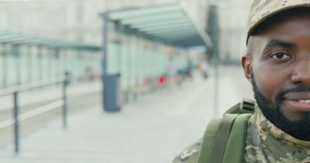 Ritratto di gioioso giovane soldato afroamericano in berretto con zaino sorridente per strada. Primo piano di mezza faccia di militare maschile con sorriso felice all'aperto alla fermata dell'autobus. Uniforme militare. — Video Stock