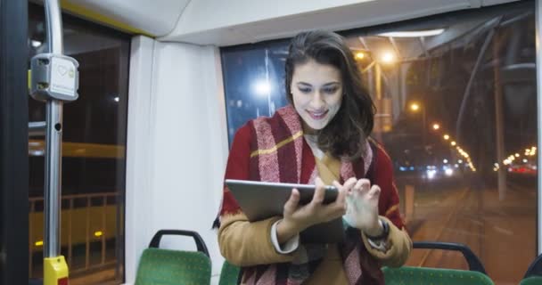 Gece eve gelip tablet aygıt kullanırken tramvayda duran paltolu ve eşarplı hoş beyaz bir kadın. Güzel bir kadın otobüsün bilgisayarda tıklaması ve kaydırması.. — Stok video