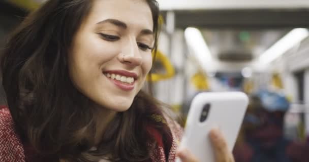 Detailní záběr běloška hezká mladá žena textové zprávy na smartphone a usmívá se, zatímco stojí v autobuse nebo tramvaji. Radostné ženské osobní rolování, ťukání a chatování na mobilním telefonu v dopravě. — Stock video