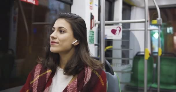 Wanita muda kulit putih bersyal dengan headphone bluetooth duduk di bus kosong di malam hari dan mendengarkan musik. femlae passanger menarik datang kembali ke rumah di trem di malam hari. — Stok Video