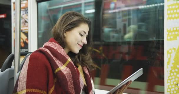 Kaukasia tersenyum pada wanita cantik dengan syal duduk di jendela di bus dan mengetuk atau menggulung pada perangkat tablet. Passanger perempuan yang menarik bergaya menggunakan komputer saat pulang ke rumah dengan trem. — Stok Video