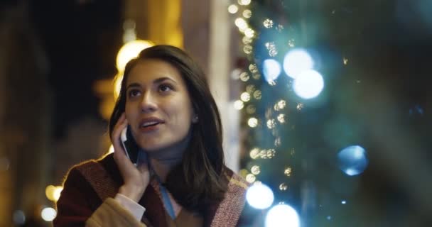 Hübsche junge kaukasische Brünette im Mantel, die spätabends vor der Tür steht und mit dem Handy telefoniert. Attraktive Frau spricht nachts mit Handy. — Stockvideo