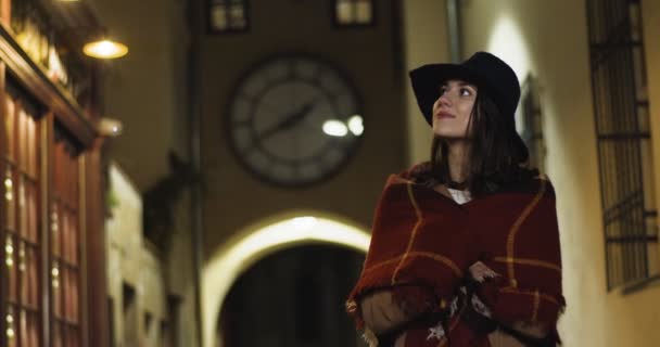 Portrait photo de belle jeune femme blanche élégante en manteau, écharpe et chapeau debout en plein air dans la cour confortable de la ville européenne avec une grande horloge sur le fond et en regardant les côtés Touriste féminine la nuit — Video