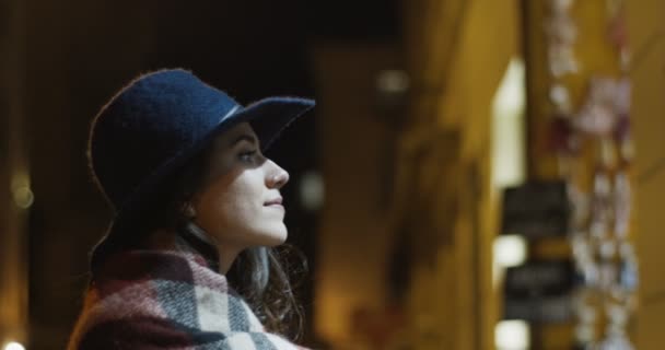 Зіткнутися з кавказькою привабливою жінкою в капелюсі, що стоїть на вулиці і дивиться на вітрину магазину вночі з посмішкою. Самка перед вітриною пізно ввечері.. — стокове відео