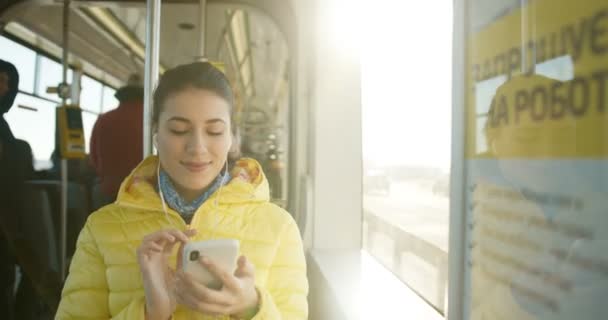 Muito jovem menina branca elegante sentado em ônibus na janela e mensagem de texto no smartphone no dia ensolarado. Mulher sorridente atraente em fones de ouvido ouvindo música no telefone em bonde de manhã . — Vídeo de Stock