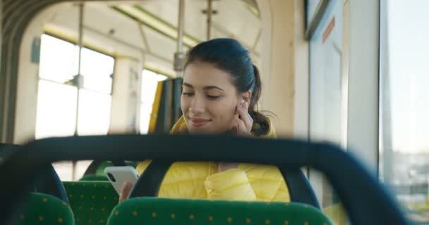 Młoda, stylowa biała dziewczyna siedząca w autobusie przy oknie i pisząca SMS-y na smartfonie w słoneczny dzień. Bardzo szczęśliwa kobieta w słuchawkach bluetooth słuchając muzyki przez telefon w pustym tramwaju rano. — Wideo stockowe