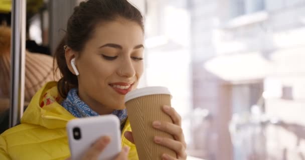 Close up de jovem caucasiana linda mulher em fones de ouvido bluetooth ouvir música no smartphone e saborear bebida quente, sentado na janela de eléctrico ou autocarro. Café feminino no transporte público . — Vídeo de Stock