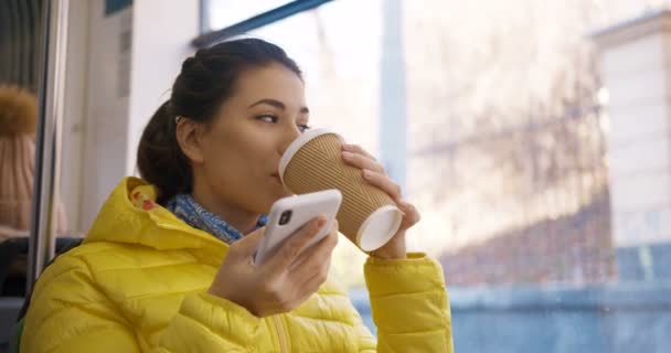 Bela jovem caucasiana sentada no bonde à janela durante o dia, tomando café para ir e gravando mensagem de voz no smartphone. Menina atraente beber bebida quente e envio de áudio no telefone . — Vídeo de Stock