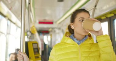 Bluetooth kulaklıklı, akıllı telefondan müzik dinleyen ve sıcak bir şeyler yudumlayan mutlu, genç, beyaz bir kız. Toplu taşımacılığın neşeli bayan yolcusu. Kahve içiyor..