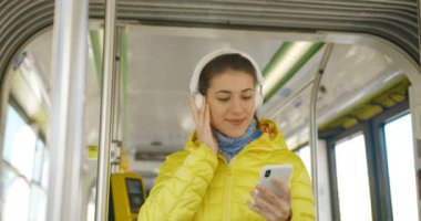 Büyük beyaz kulaklıklı neşeli beyaz bir kız akıllı telefondan müzik dinliyor ve tramvayda gülümsüyor. Telefondaki oyuncuyu kullanarak toplu taşımanın neşeli kadın yolcusu.
