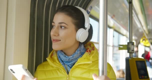 Närbild av glada glada glada unga kaukasiska söt flicka i stora vita hörlurar lyssnar på musik på smartphone och leende. Glädjande kvinnlig passagerare kollektivtrafik med hjälp av spelare på telefon. — Stockvideo