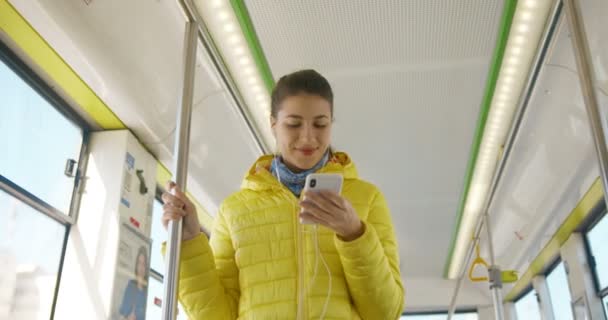 Veja de baixo em caucasiano jovem menina muito elegante de pé em bonde durante o dia e usando smartphone para ouvir música em fones de ouvido. feminino passanger de transporte público ouvindo música . — Vídeo de Stock