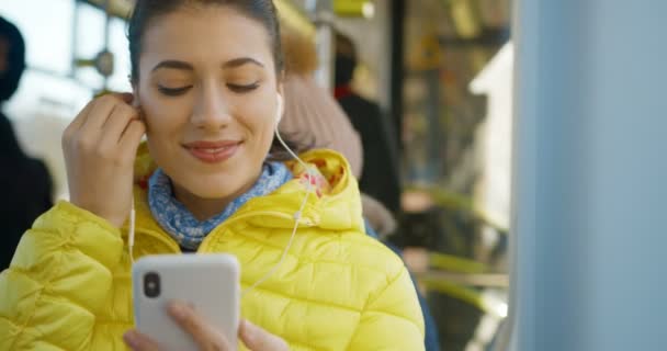 Sarı montlu, otobüste oturan ve kulaklarına kulaklık takan güzel beyaz bir kadın. Mutlu kız sabah toplu taşıma aracıyla giderken akıllı telefondan müzik dinliyor.. — Stok video