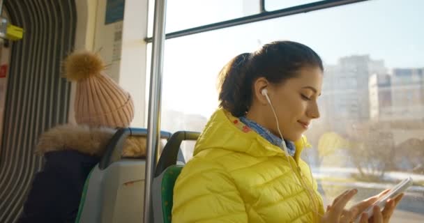 Glada unga kaukasiska söt flicka i bluetooth hörlurar lyssna på musik på smartphone och leende. Glädjande glad kvinnlig passagerare kollektivtrafik med hjälp av spelare på telefon. — Stockvideo