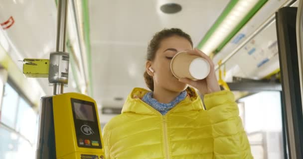 Vrolijk vrolijk jong Kaukasisch mooi meisje in bluetooth koptelefoon luisteren naar muziek op de smartphone en nippen warme drank. Vrolijke vrouwelijke passagier van het openbaar vervoer drinken koffie to-go. — Stockvideo