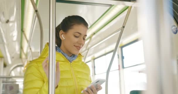 Ευτυχισμένη χαρούμενη νεαρή Καυκάσια όμορφη κοπέλα με ακουστικά bluetooth ακούγοντας μουσική στο smartphone και χαμογελώντας στο τραμ. Χαρούμενη γυναίκα passanger των μέσων μαζικής μεταφοράς χρησιμοποιώντας παίκτη στο τηλέφωνο. — Αρχείο Βίντεο