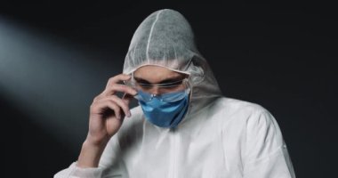 Tam teçhizatlı kostüm ve tıbbi maske takmış beyaz erkek doktor gözlük takıyor ve arka plandaki siyah duvara bakıyor. Küresel salgın konsepti. Doktor epidemiyoloji uzmanı için koruma.