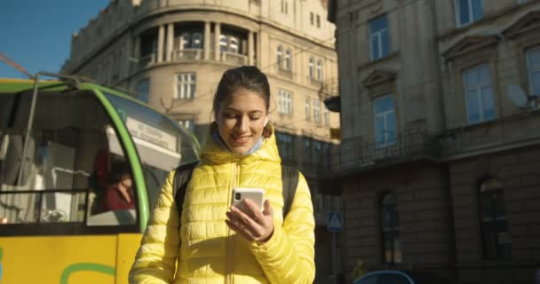 Κομψό νεαρό καυκάσιο κορίτσι με ακουστικά bluetooth στέκεται σε στάση αστικών μεταφορών και γραπτών μηνυμάτων στο smartphone. Όμορφη θηλυκή υποκλοπή και κύλιση στο κινητό τηλέφωνο εξωτερική. Τραμ που διέρχεται πίσω — Αρχείο Βίντεο