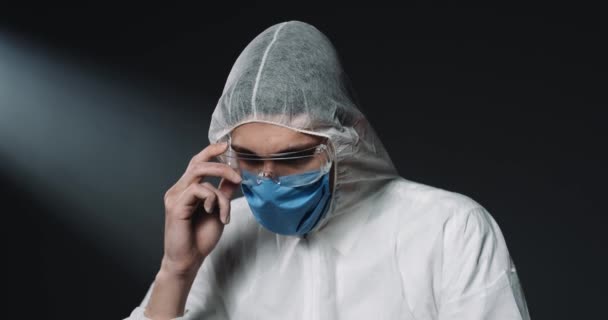 Портрет молодого врача-кавказца в полностью оборудованном костюме и медицинской маске, надевающего очки и смотрящего на камеру на тёмном фоне. Глобальная пандемия. Защита для эпидемиолога . — стоковое видео