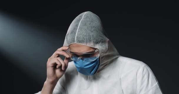 Портрет уставшего кавказца молодого медика в полностью оборудованном костюме и медицинской маске, надевающего очки и смотрящего на камеру на темном фоне. Глобальная пандемия концепции ковида-19 Лабораторный работник — стоковое видео